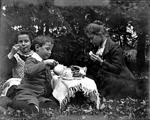 Photo of two girls and a boy taking tea outdoors in old fashioned clohting