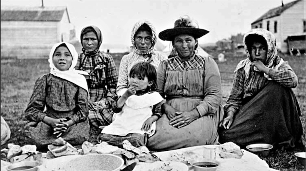 Two Indigenous women having a picnic with four children. They're all wearing bonnets and fine dresses.